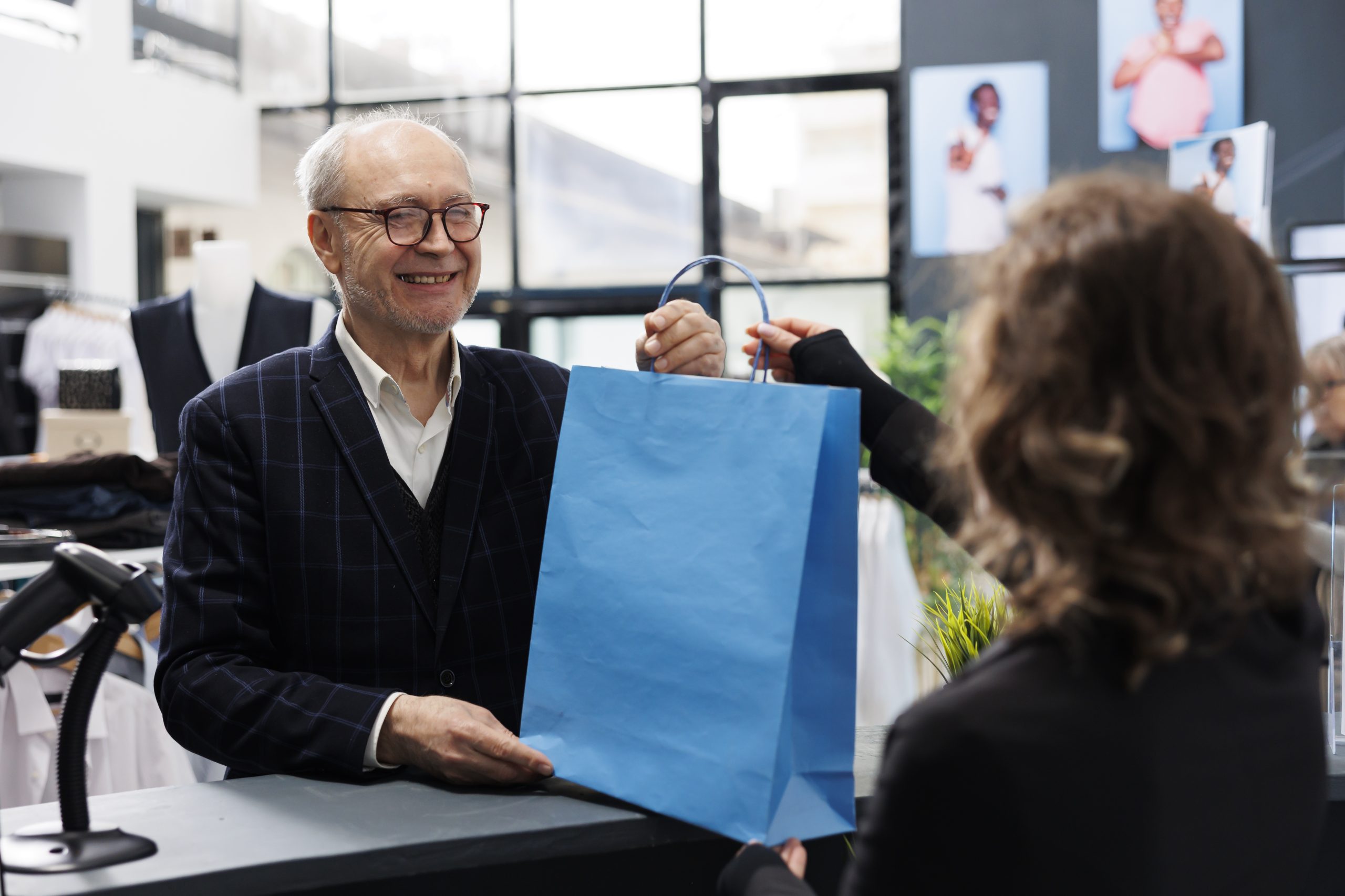 Elderly shopaholic man buying elegant clothes in retail store, making electronic transaction at counter desk. Cheerful customer talking to store employee discussing promotions, commercial activity.